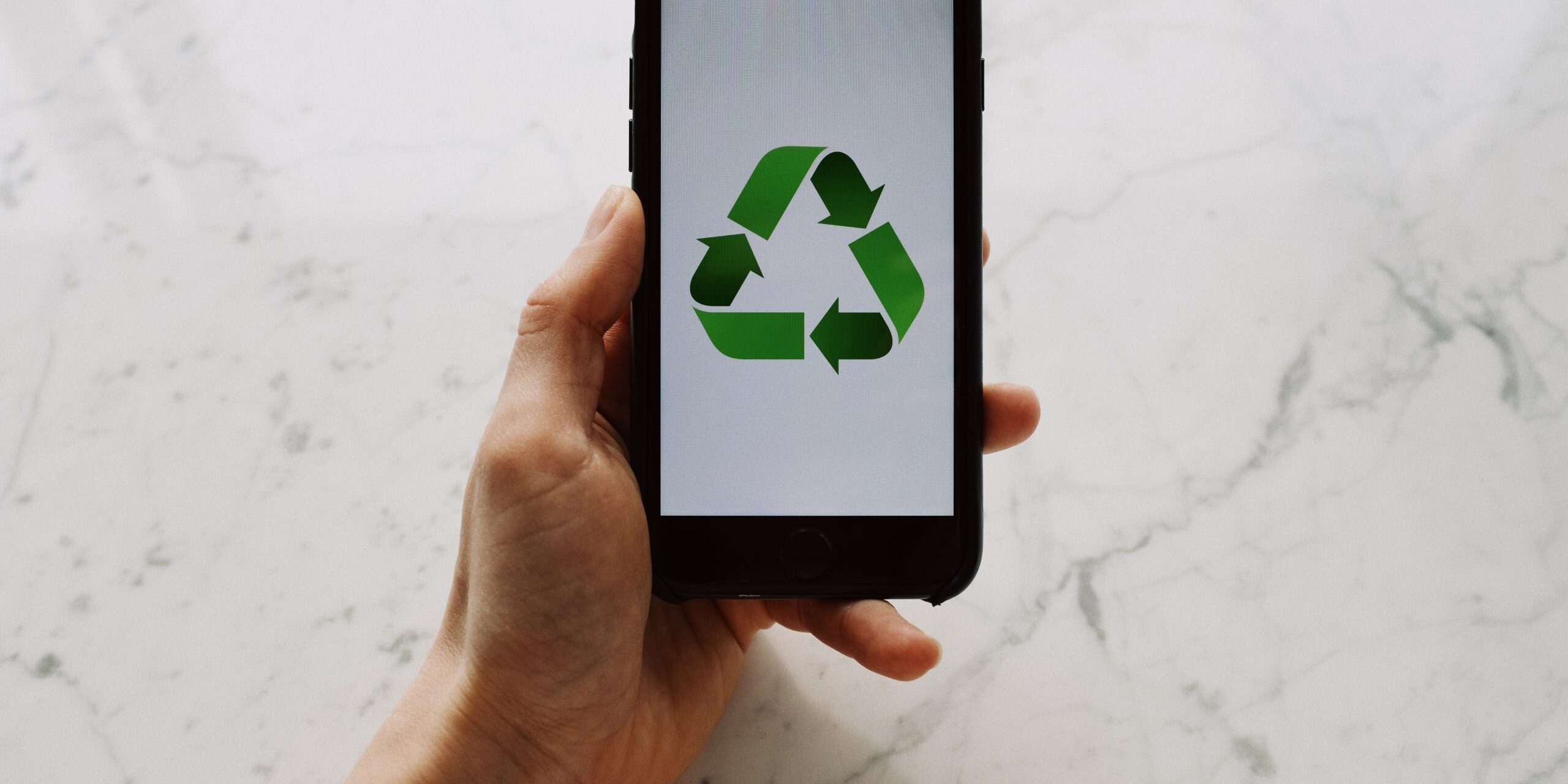Important-Steps-to-Take-Before-You-Recycle-image-scaled.jpg From above view of faceless person holding mobile phone with white screen and green recycle logo above marble surface
